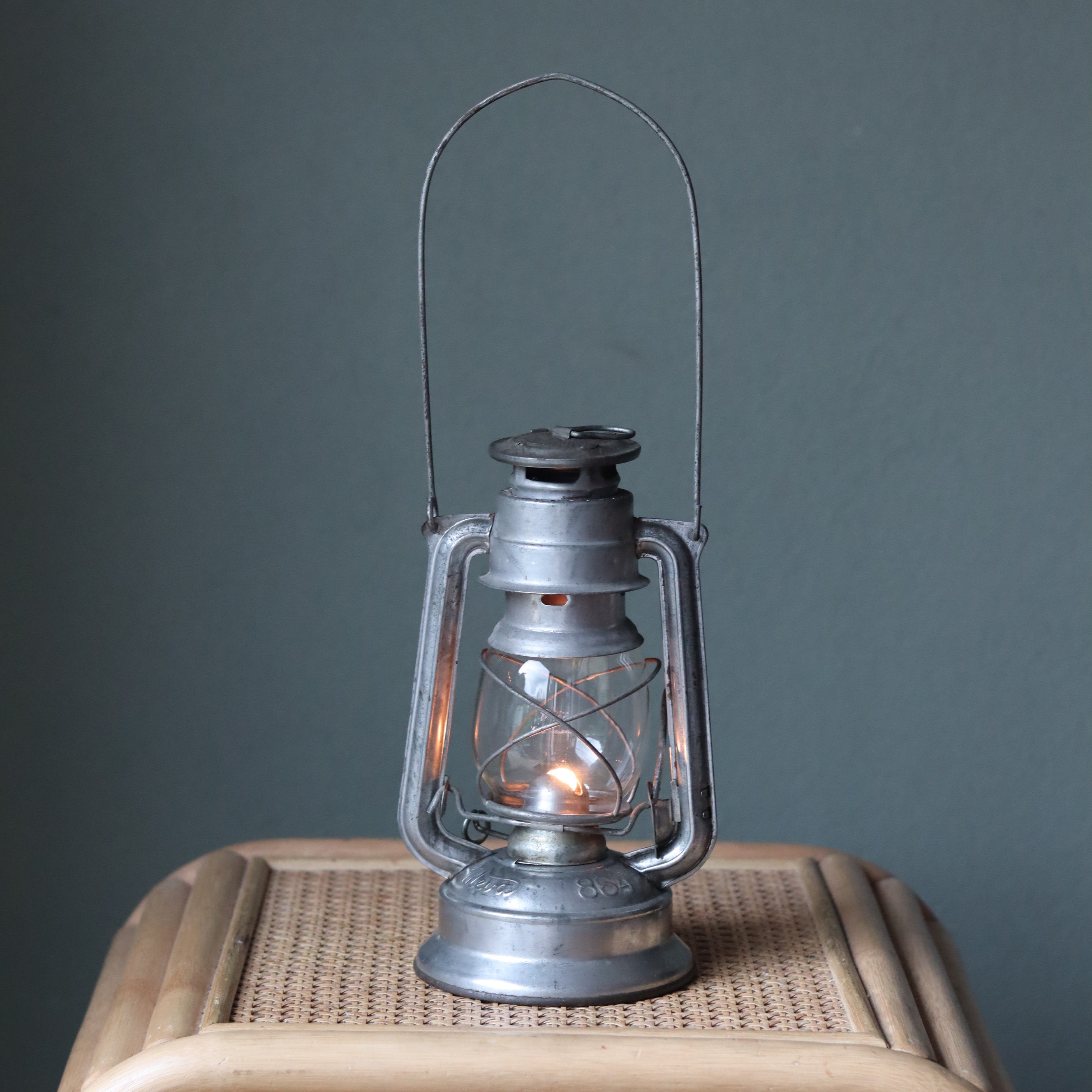 Vintage Mewa Hurricane Lantern – Model 864 (1978) oil lamp with glowing flame and classic metal design on wicker surface