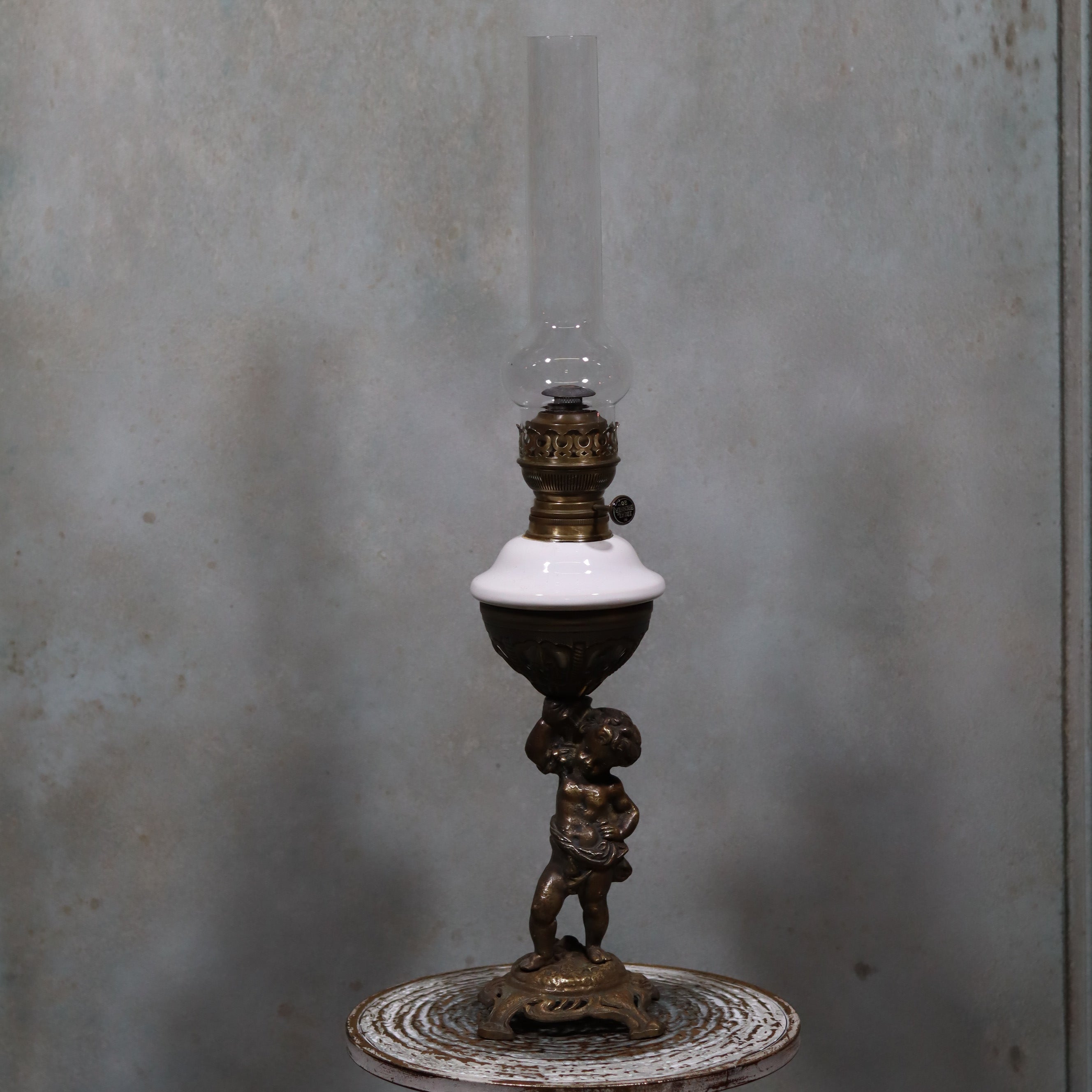 Vintage XXL Oil Lamp with BJB Matador Burner – Bronze Base with Cherub Design standing on a rustic round table against a gray wall.